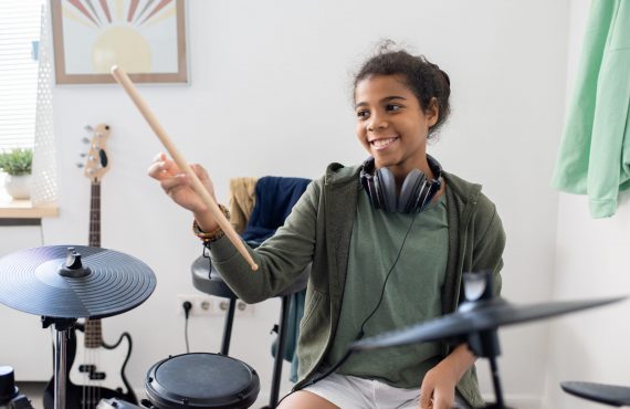 Happy cute biracial schoolgirl with drumsticks hitting drums at lesson of music in home environment